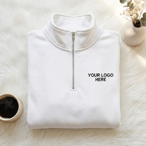 Può includere: Una felpa bianca con zip a un quarto e collo alto con una cerniera in metallo color argento. L'area del petto presenta il testo nero "YOUR LOGO HERE" ricamato sul lato destro del capo, che è posizionato in piano.