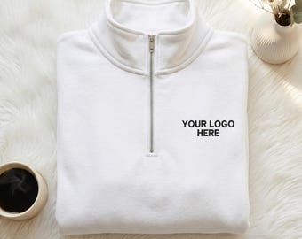May include: A white quarter-zip sweatshirt featuring a high collar and a silver-toned metal zipper. The chest area displays the black embroidered text "YOUR LOGO HERE" positioned to the right of the center zipper closure.