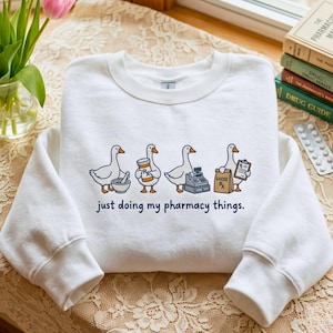 May include: A white crewneck sweatshirt featuring a graphic of four geese performing pharmacy tasks, including holding a mortar and pestle, a pill bottle, a cash register, and a prescription bag. The text reads, "just doing my pharmacy things."