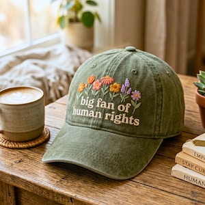 Puede incluir: Gorra de béisbol verde oliva con flores bordadas y el texto "big fan of human rights". La gorra presenta una visera curva y un diseño clásico de seis paneles. El bordado floral incluye flores naranjas, rosas y moradas.