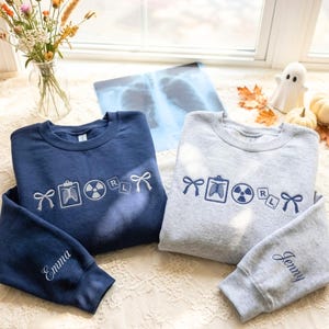 Peut inclure: Un sweat-shirt bleu marine et un sweat-shirt gris chiné, chacun orné de broderies représentant un nœud, une radiographie thoracique sur un presse-papier, un symbole de radiation et les lettres R et L. La manche marine porte le nom Emma, la grise Jenny.