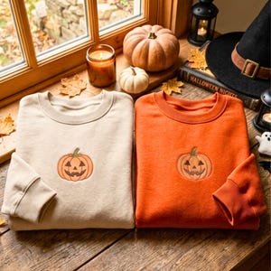 Op de afbeelding: Twee sweaters met ronde hals, één in beige en één in oranje, elk voorzien van een kleine geborduurde jack-o'-lantern pompoen op het midden van de borst. De sweaters hebben lange mouwen met geribbelde boorden en een ronde hals.