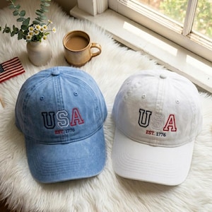 Op de afbeelding: Twee baseballpetten, één in lichtblauw denim en één in wit, beide voorzien van geborduurde tekst met USA in grote letters en EST. 1776 eronder. De petten hebben een gestructureerde kroon, een gebogen klep en een knoopje bovenop.