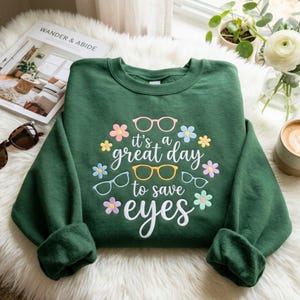 May include: A dark green crewneck sweatshirt featuring a graphic print with the text "it's a great day to save eyes" in white script. The design includes illustrations of three pairs of eyeglasses in pink, yellow, and light blue, surrounded by small colorful flowers.