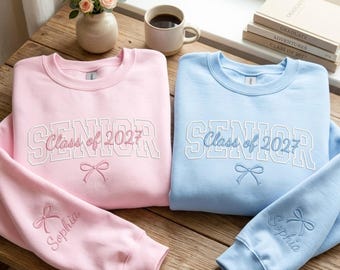 Op de afbeelding: Twee sweaters met ronde hals in lichtroze en lichtblauw. Beide hebben wit geborduurde tekst SENIOR, cursieve tekst Class of 2027 en een strikicoon. De mouwen zijn geborduurd met een strik en de naam Sophia in bijpassende kleuren.