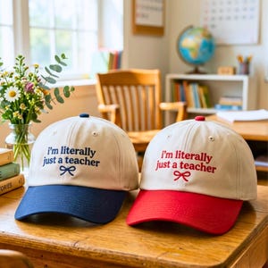Pode incluir: Dois bonés de beisebol com copas na cor creme e abas curvas. Um boné tem aba azul-marinho e bordado azul-marinho com a frase 'i'm literally just a teacher' e um pequeno laço. O outro tem aba vermelha e bordado vermelho com o mesmo texto.