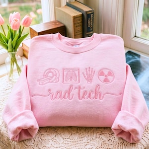 May include: A light pink crewneck sweatshirt featuring embroidered designs of an MRI machine, a rib cage, a hand skeleton, and a radiation symbol. Below these icons, the text rad tech is embroidered in a cursive script font across the chest.