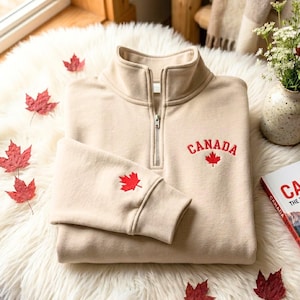 May include: A beige quarter-zip sweatshirt featuring the word CANADA embroidered in red thread above a red maple leaf on the chest, with an additional red maple leaf embroidered on the sleeve cuff and a silver-toned metal zipper pull.