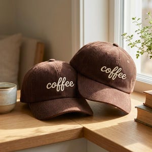 May include: Two brown corduroy baseball caps, each embroidered with the word "coffee" in white script. The caps sit on a wooden surface. The caps are a warm brown colour, suggesting a casual style. The caps are a warm brown colour.