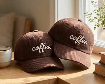 May include: Two brown corduroy baseball caps with the word "coffee" embroidered in white script. The caps are resting on a wooden surface, suggesting a casual, everyday style. The caps are a warm brown color.