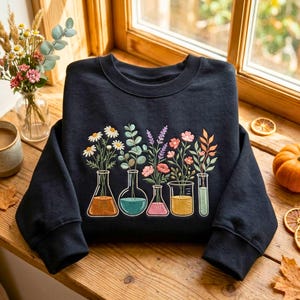 Könnte beinhalten: Ein schwarzes Sweatshirt mit Rundhalsausschnitt und einer Stickerei von fünf Laborgefäßen aus Glas, darunter Kolben und Bechergläser, die jeweils verschiedene bunte Wildblumen und Grünpflanzen mit orangefarbenen, blauen, rosafarbenen und gelben Flüssigkeiten enthalten.