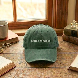 May include: A green baseball cap with the text "coffee & books" in white. The hat is sitting on a wooden surface near a window, with books and a coffee cup in the background. The cap has a curved brim.