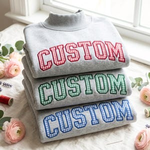 May include: A stack of three light grey mock neck sweatshirts, each featuring the word CUSTOM embroidered in a collegiate font. The top sweatshirt has red gingham letters, the middle has green gingham, and the bottom has blue gingham.