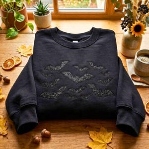 May include: A black long-sleeve crewneck sweatshirt featuring a central embroidered pattern of multiple flying bats. The bats are rendered in a dark, shimmering, glittery thread that contrasts against the solid black fabric of the garment.