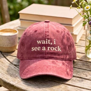 May include: A maroon baseball cap with a distressed finish featuring the embroidered white text "wait, i see a rock" centered on the front panel. The cap has a curved brim and a structured crown with ventilation eyelets on the top panels.