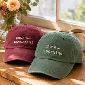 May include: A maroon baseball cap and a sage green baseball cap, both featuring the embroidered white text "pause... nevermind" on the front panels. Each cap has a curved brim, a button on top, and stitched eyelets for ventilation.