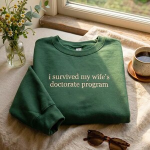 May include: A dark green sweatshirt with the text "i survived my wife's doctorate program" in white. The sweatshirt is folded on a light-colored surface, with a pair of sunglasses and a cup of coffee nearby. A vase of flowers is also visible.
