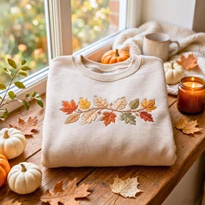 Könnte beinhalten: Ein beigefarbenes Sweatshirt mit Rundhalsausschnitt und einem horizontalen Stickmotiv aus Herbstblättern in Orange-, Gelb-, Braun- und Grüntönen auf der Brustmitte. Der Stoff ist glatt und der Halsausschnitt ist gerippt.