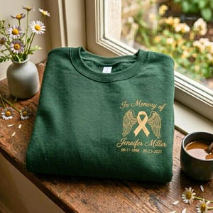 May include: A dark green crewneck sweatshirt featuring a gold-colored graphic on the chest. The design includes a ribbon with angel wings, the text In Memory of Jennifer Miller, and the dates 09-11-1998 - 05-23-2025 printed in gold ink.