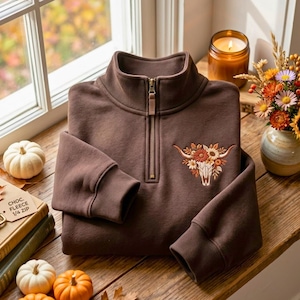 May include: A dark brown quarter-zip pullover sweatshirt featuring an embroidered design on the chest of a white cow skull adorned with a colorful arrangement of orange, yellow, and white flowers and green leaves.