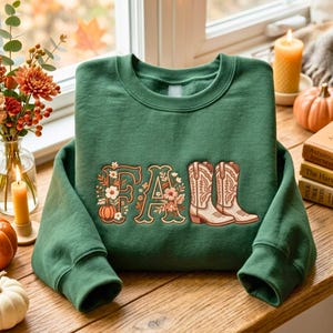 May include: A forest green crewneck sweatshirt featuring a graphic design with the word FALL in a decorative font. The letter F is adorned with floral illustrations and a small pumpkin, while the letter L is replaced by two tan cowboy boots.