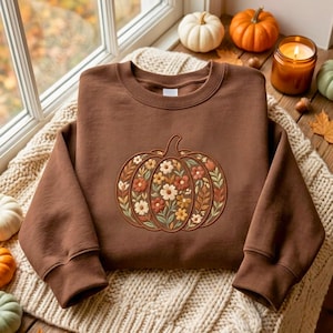 May include: A chocolate brown crewneck sweatshirt featuring a central embroidered pumpkin design. The pumpkin is filled with a detailed pattern of small white, orange, and yellow flowers surrounded by green leaves and thin brown stems.
