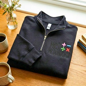 May include: A black quarter-zip sweatshirt with the name "MRS. MCDONOUGH" embroidered in black thread. Colourful maths symbols are embroidered next to the name. The sweatshirt has a gold zip and a ribbed cuff.