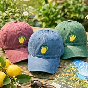 May include: Three baseball caps in shades of red, blue, and green, each embroidered with a yellow lemon and green leaves. The caps are displayed on a wooden surface with lemons and a travel magazine.