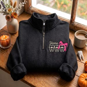 May include: A black quarter-zip sweatshirt featuring an embroidered design on the left chest. The design includes the name Stevie in white script, three white teeth with metal braces, and a bright pink bow tied around the teeth.
