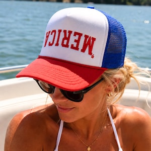 Könnte beinhalten: Eine rot-weiß-blaue Trucker-Mütze mit dem Wort "MERICA" in roten Blockbuchstaben. Die Mütze hat einen roten Schirm, eine weiße Vorderseite und eine blaue Mesh-Rückseite. Die Mütze wird von einer Person mit Sonnenbrille getragen.