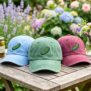 May include: Three baseball caps in light blue, sage green, and muted red, each featuring an embroidered green pickle design on the front panel. The caps have a structured crown, a curved brim, and a washed fabric texture with visible stitching.