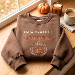May include: A brown crewneck sweatshirt featuring the embroidered text GROWING A LITTLE Pumpkin in white and orange thread. Below the text is an outline of a pumpkin with two small orange baby footprints centred inside the pumpkin design.