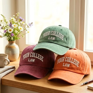 Può includere: Tre cappellini da baseball verdi, bordeaux e arancioni sono impilati su una superficie di legno. Ogni cappellino presenta la scritta bianca "YOUR COLLEGE LAW". Sullo sfondo un vaso di fiori e una tazza.