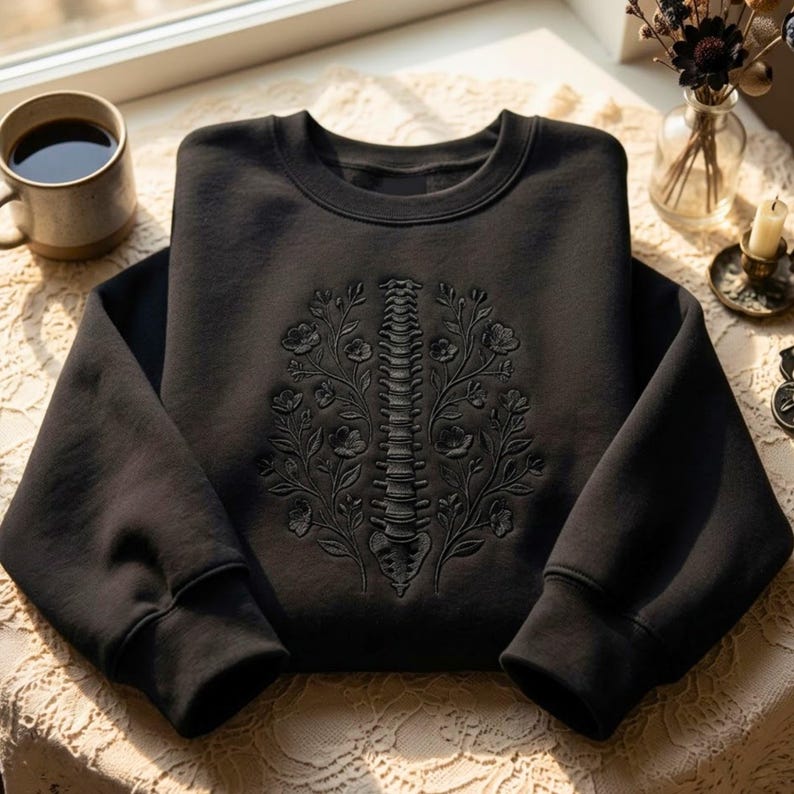 Op de afbeelding: Een zwart sweatshirt met ronde hals en een ton-sur-ton borduursel op de borst. Het ontwerp toont een menselijke ruggengraat in het midden tussen twee symmetrische bloemtakken met bladeren en bloesems, volledig uitgevoerd in zwart garen.