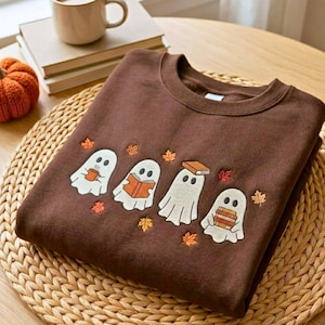 Puede incluir: Una sudadera de cuello redondo de color marrón oscuro con un diseño bordado de cuatro fantasmas blancos. Un fantasma sostiene una taza, dos sostienen libros y uno tiene un libro sobre la cabeza. El diseño incluye pequeñas hojas de otoño bordadas.