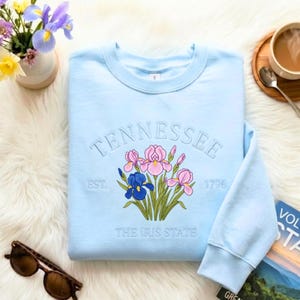 Op de afbeelding: Een lichtblauw sweatshirt met ronde hals en een geborduurd ontwerp van paarse en blauwe irissen. De tekst "TENNESSEE", "EST. 1796" en "THE IRIS STATE" is in bijpassend lichtblauw garen rond de bloemenafbeelding geborduurd.