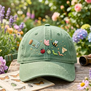 May include: A sage green baseball cap featuring embroidered insects and a glass jar. The embroidery includes a ladybug, a bee, a caterpillar, a moth, a butterfly, and a small jar with a cork stopper, accented with tiny yellow stars and dotted lines.
