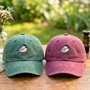 Puede incluir: Dos gorras de béisbol, una en verde apagado y otra en granate profundo, cada una con un diseño bordado de un pequeño pájaro posado en una rama de pino. Las gorras tienen una textura suave y lavada con una visera curva.