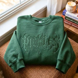 Könnte beinhalten: Ein waldgrünes Sweatshirt mit Rundhalsausschnitt und dem Wort Pickles, das in einer dunkelgrünen gotischen Schrift auf der Brust eingestickt ist. Das Kleidungsstück hat lange Ärmel mit gerippten Bündchen und einen gerippten Halsausschnitt.