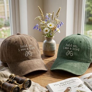 May include: Two baseball caps, one brown and one green, are displayed on a wooden surface. Each cap has the embroidered text "hold on, i see a bird" with binoculars. A vase of flowers and a birdwatching guide are also present.