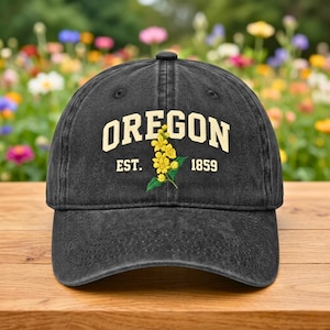 May include: A black baseball cap with the word "OREGON" in cream-colored block letters. Below the word "OREGON" is the text "EST. 1859" and a yellow flower with green leaves. The cap is sitting on a wooden surface.