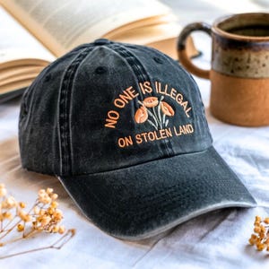 May include: Black baseball cap with the embroidered text "NO ONE IS ILLEGAL ON STOLEN LAND" in orange. The cap features an orange floral design and a curved brim. The hat is sitting on a white surface.
