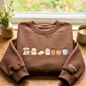 May include: A brown crewneck sweatshirt featuring an embroidered line of baking ingredients including a mixer, flour bag, butter, sugar, cocoa, eggs, and a measuring cup. The name Emma is embroidered in white cursive on the right sleeve cuff.