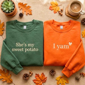 May include: A dark green sweatshirt with the text She's my sweet potato and an orange sweatshirt with the text I yam and a small heart icon. Both garments feature long sleeves and crew necklines, displayed with autumn leaves and a coffee mug.