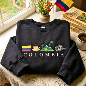 Può includere: Una felpa nera a girocollo con un ricamo raffigurante la bandiera colombiana, una tazza di caffè fumante, montagne verdi con palme e un cappello tradizionale, con la parola COLOMBIA ricamata in lettere maiuscole bianche sottostanti.