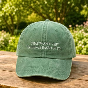 May include: A sage green baseball cap with a six-panel construction and a curved brim. The front features white embroidered text that reads, "THAT WASN'T VERY EVIDENCE BASED OF YOU," centered in a clean, sans-serif font across the crown.