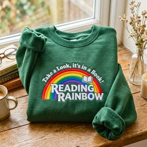 May include: A forest green crewneck sweatshirt featuring an embroidered graphic of a colorful rainbow and an open book. The text reads Take a Look, it's in a Book! and READING RAINBOW in white lettering centered on the chest of the garment.