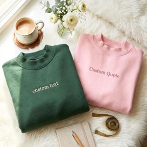 Puede incluir: Una sudadera verde oscuro con el texto bordado custom text y una sudadera rosa claro con el texto bordado Custom Quote. Ambas prendas cuentan con cuello redondo acanalado y mangas largas, presentadas como un par de prendas a juego.