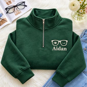 May include: A dark green quarter-zip sweatshirt featuring an embroidered design on the left chest. The embroidery depicts a pair of white glasses with one eye open and one winking, positioned above the name Aidan in white lettering.