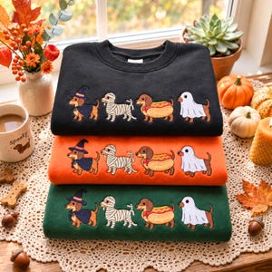 May include: A stack of three crewneck sweatshirts in black, orange, and dark green. Each sweatshirt features an identical embroidered design of four dachshunds dressed for Halloween: one as a witch, one as a mummy, one as a hot dog, and one as a ghost.